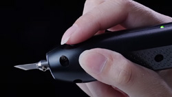 A closeup of an ultrasonic cutter held by a person. It is black, about the size and shape of an electric toothbrush. It has an exacto type blade at the end.