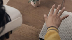 A view from behind and to the right of a person wearing a white VR headset holdng their hand above a white, flat device with small holes covering the top.