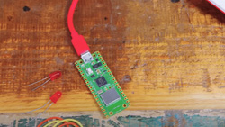 A small circuit board is connected to a laptop via a red USB cable. Two unsoldered red LEDs sit next to it.